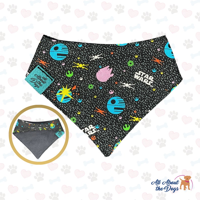 May include: Black bandana with a Star Wars themed print featuring spaceships, planets, and the words "Star Wars". The bandana is made of cotton and has a grey underside. The bandana is tied around a dog's neck.