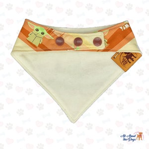 May include: A dog bandana with a cream-colored triangle base and a top section featuring a repeating pattern of a cartoon character and orange stripes. The bandana has three brown buttons and a small leather tag.