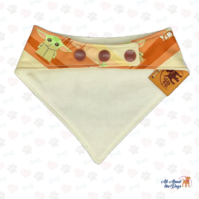 May include: A dog bandana with a cream-colored triangle base and a top section featuring a repeating pattern of a cartoon character and orange stripes. The bandana has three brown buttons and a small leather tag.