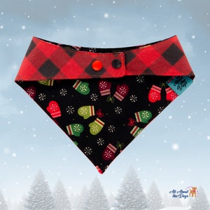 May include: A festive dog bandana with a red and black buffalo plaid top and a black bottom patterned with green and red mittens and white snowflakes. The bandana has two snap closures.