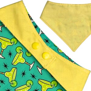May include: A pet bandana with a turquoise background featuring margarita glasses and star patterns. The bandana has a yellow trim and two yellow snap closures. A separate yellow bandana is also visible.