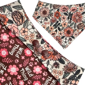 May include: Two dog bandanas with floral patterns. One bandana is brown with pink flowers and the words "Wild Thing". The other bandana is cream-colored with pink and orange flowers. Both bandanas have a snap closure.