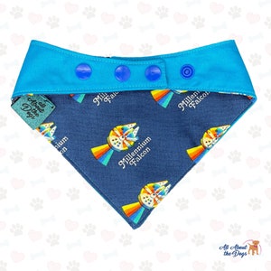 May include: A blue and navy blue dog bandana with a repeating pattern of the Millennium Falcon spaceship with a rainbow behind it. The bandana has a blue snap closure and the text "All About the Dogs" is on the corner.