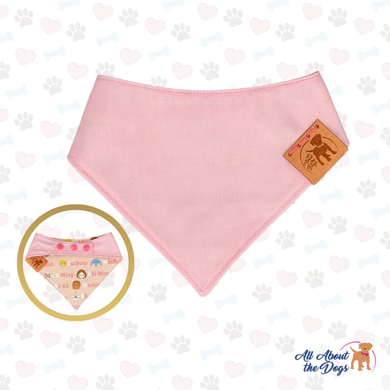 May include: A light pink dog bandana with a stitched edge and a brown leather tag. The bandana is triangular and has a smaller, patterned bandana in a gold circle. The smaller bandana has text and cartoon characters.