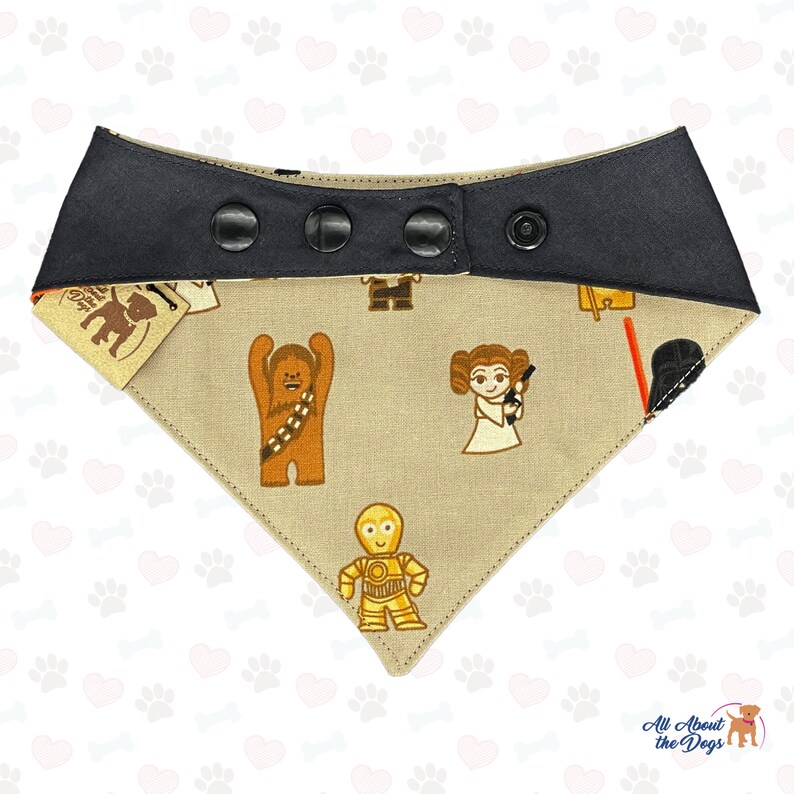 K&ouml;nnte beinhalten: Ein beiger Hundehalstuch mit schwarzem Besatz und Druckkn&ouml;pfen. Das Halstuch zeigt ein sich wiederholendes Muster von Star Wars-Charakteren, darunter Chewbacca, Prinzessin Leia und C-3PO.