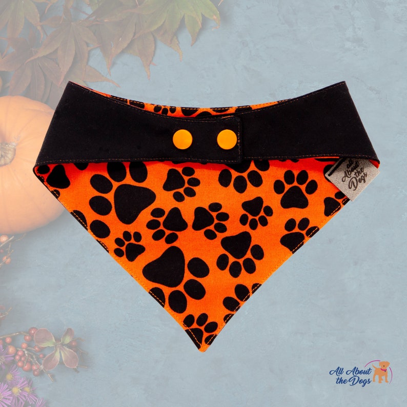 Peut inclure: Bandana pour chien orange et noir sur le th&egrave;me d'Halloween. Le bandana pr&eacute;sente des empreintes de pattes noires sur un fond orange. La bande sup&eacute;rieure est noire avec deux boutons orange. L'&eacute;tiquette indique "All About the Dogs".