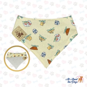 May include: A beige bandana with a repeating pattern of a green alien character, a spaceship, and a helmet. The bandana has a snap closure and is made of cotton.