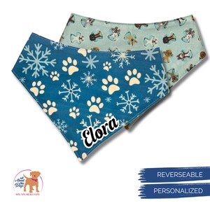 K&ouml;nnte beinhalten: Zwei wendbare Hunde-Bandanas. Eines ist blau mit wei&szlig;en Schneeflocken und Pfotenabdr&uuml;cken, das andere hellblau mit Hundeillustrationen. Auf dem blauen Bandana steht der Name "Elora". Die Bandanas sind personalisiert.