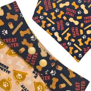 May include: Close-up of two dog bandanas. One is light tan with paw prints and the words "BONE PETIT". The other is navy blue with bone and treat graphics and the words "TREAT TASTER".
