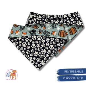 May include: Reversible dog bandana. One side features a black and white paw print pattern, the other a light blue background with bourbon barrel and "Bourbon Buddy" text. Includes snap closures.