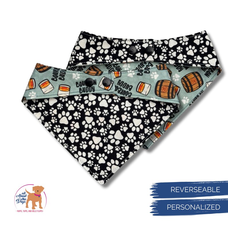May include: Reversible dog bandana. One side features a black and white paw print pattern, the other a light blue background with bourbon barrel and "Bourbon Buddy" text. Includes snap closures.