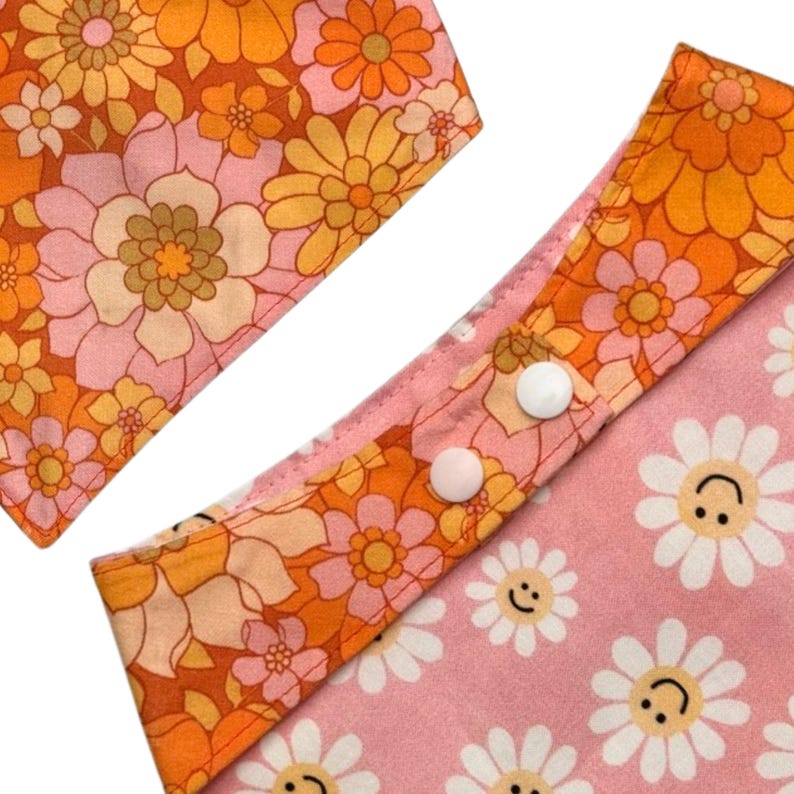Puede incluir: Dos piezas de tela con un estampado floral en naranja, amarillo y rosa. Una pieza es un babero con broches blancos y un fondo rosa con caras de margaritas sonrientes. La otra pieza es cuadrada.