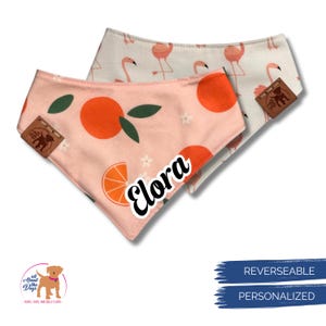 May include: Two reversible dog bandanas. One is peach with orange fruit and green leaves, the other is white with pink flamingos. The peach bandana has the name "Elora" in white script. Both have brown leather tags.