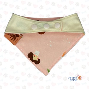 Peut inclure: Bandana rose pour chien avec un col cr&egrave;me et trois boutons-pression blancs. Le bandana pr&eacute;sente un motif r&eacute;p&eacute;t&eacute; d'un personnage de dessin anim&eacute; et d'&eacute;toiles. Une &eacute;tiquette marron avec un logo est attach&eacute;e.