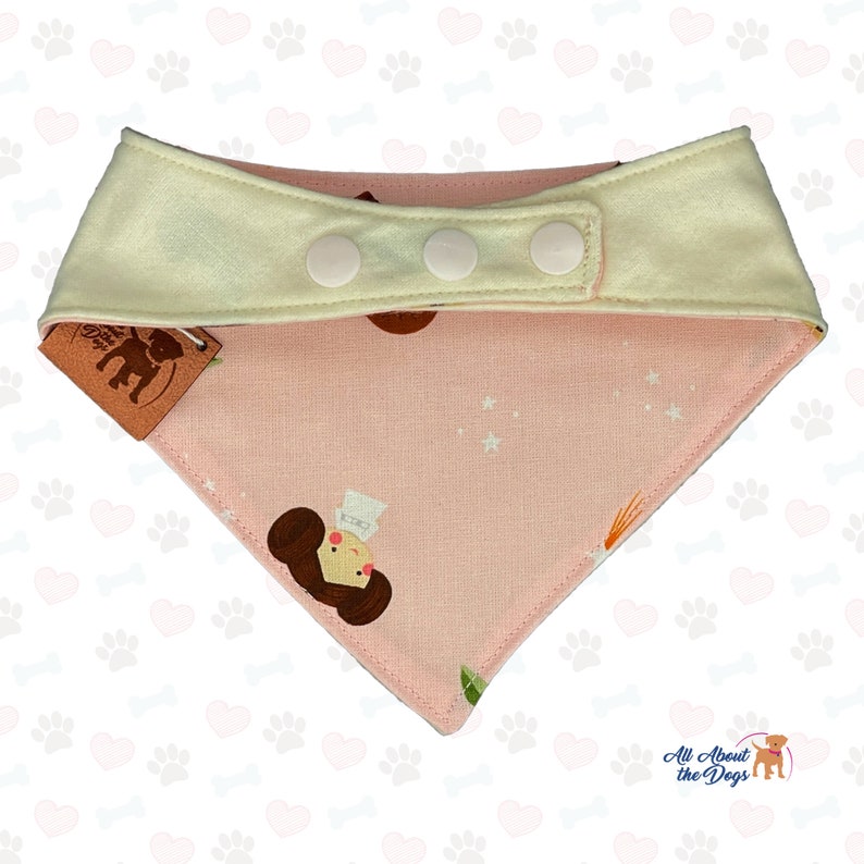 Peut inclure: Bandana rose pour chien avec un col cr&egrave;me et trois boutons-pression blancs. Le bandana pr&eacute;sente un motif r&eacute;p&eacute;t&eacute; d'un personnage de dessin anim&eacute; et d'&eacute;toiles. Une &eacute;tiquette marron avec un logo est attach&eacute;e.