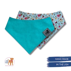 Pu&ograve; includere: Due bandane per cani. Una &egrave; turchese tinta unita, l'altra ha uno sfondo azzurro con fiori rosa e calici di vino. Entrambe le bandane hanno una piccola etichetta con una silhouette di cane. Sono visibili anche le parole "HAND MADE" e "IN THE USA".