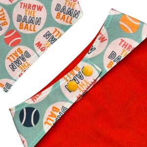 Peut inclure: Un bandana rouge pour chien avec une bordure vert d'eau clair. Il comporte le texte "THROW THE DAMN BALL" dans un cercle blanc avec un graphique de balle de tennis. Fermetures &agrave; boutons-pression jaunes.