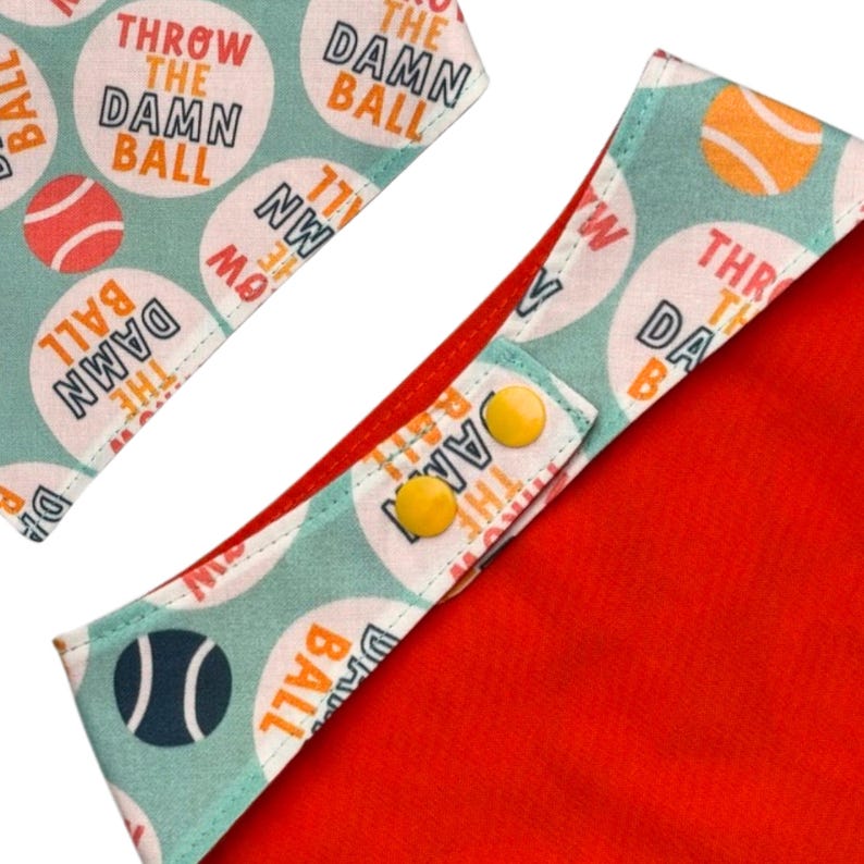 Peut inclure: Un bandana rouge pour chien avec une bordure vert d'eau clair. Il comporte le texte "THROW THE DAMN BALL" dans un cercle blanc avec un graphique de balle de tennis. Fermetures &agrave; boutons-pression jaunes.