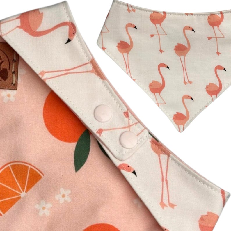 May include: Two baby bibs with a flamingo theme. One bib features a repeating pattern of pink flamingos on a white background. The other bib has a pink background with orange slices and white flowers. Both bibs have snap closures.