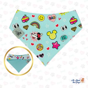 May include: A light blue dog bandana featuring cartoon graphics such as a rainbow, hearts, stars, and a cartoon character. The bandana has a folded over design and a tag with a dog silhouette.