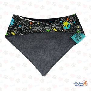 May include: Black and grey dog bandana with a colorful space-themed print and three black buttons. The bandana has a teal tag that says "All About the Dogs".