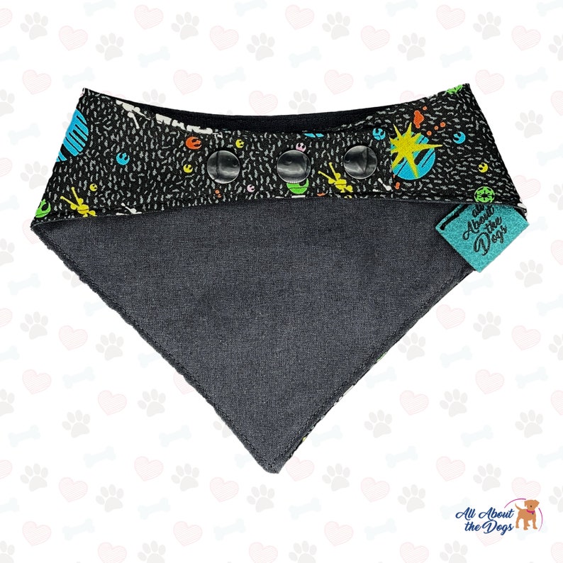 May include: Black and grey dog bandana with a colorful space-themed print and three black buttons. The bandana has a teal tag that says "All About the Dogs".