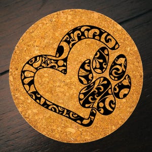 Puede incluir: Posavasos redondo de corcho con un diseño de corazón y huella de pata en negro. El corazón y la huella de la pata están llenos de intrincados patrones arremolinados. El posavasos es de color marrón claro.
