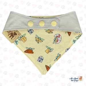 May include: A light grey dog bandana with a yellow button closure. The bandana is reversible and features a light yellow fabric with a repeating pattern of a green alien character and a Mandalorian helmet.