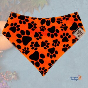 Peut inclure: Bandana orange pour chien avec des empreintes de pattes noires. Le bandana triangulaire pr&eacute;sente un motif r&eacute;p&eacute;t&eacute; d'empreintes de pattes. Une petite &eacute;tiquette indique "All About the Dogs".