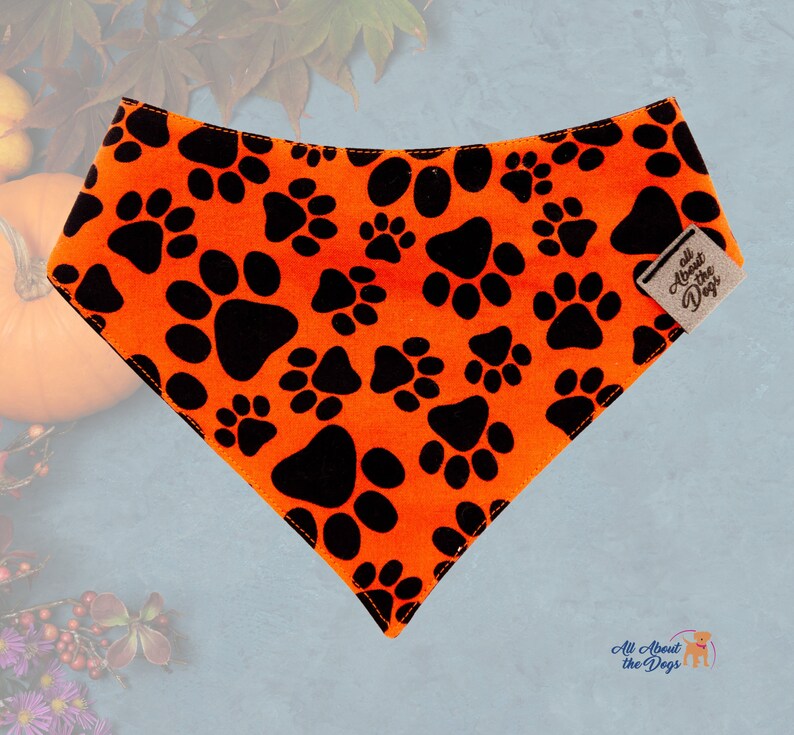 Peut inclure: Bandana orange pour chien avec des empreintes de pattes noires. Le bandana triangulaire pr&eacute;sente un motif r&eacute;p&eacute;t&eacute; d'empreintes de pattes. Une petite &eacute;tiquette indique "All About the Dogs".