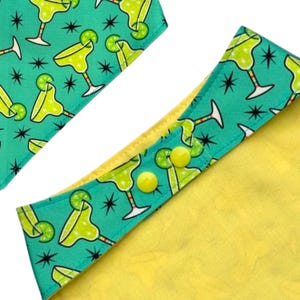 May include: Close-up of a dog bandana with a margarita cocktail pattern on a teal background. The bandana has a yellow interior and yellow buttons. The design features lime green margaritas and black starbursts.