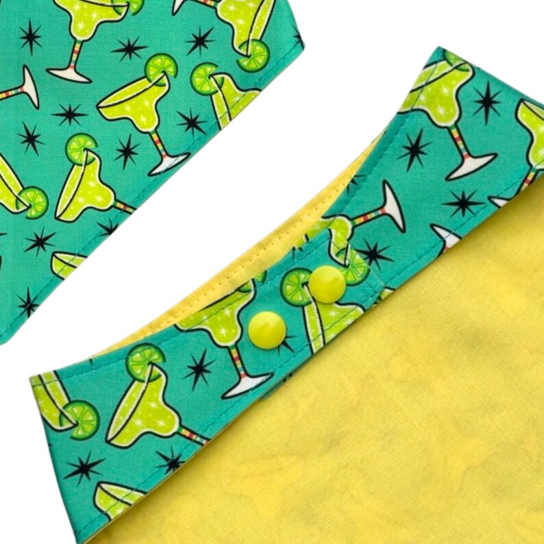 May include: Close-up of a dog bandana with a margarita cocktail pattern on a teal background. The bandana has a yellow interior and yellow buttons. The design features lime green margaritas and black starbursts.