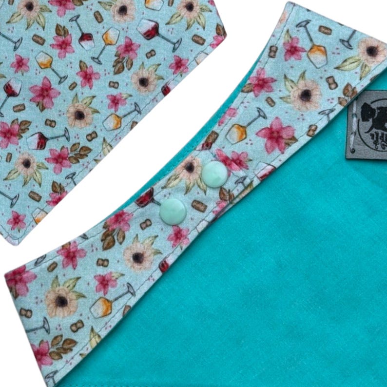 Pu&ograve; includere: Primo piano di una bandana turchese per cani con un motivo floreale e calici di vino. La bandana ha un retro turchese ed &egrave; fissata con due bottoni verde chiaro. Un accessorio abbinato &egrave; visibile in alto a sinistra.