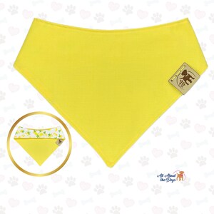 Pu&ograve; includere: Un bandana giallo brillante per cani, di forma triangolare. Una piccola immagine circolare mostra un dettaglio del bandana con un motivo a limoni bianchi e gialli. Un'etichetta marrone con un'illustrazione di un cane &egrave; attaccata. Le parole "All About the Dogs" sono in basso.