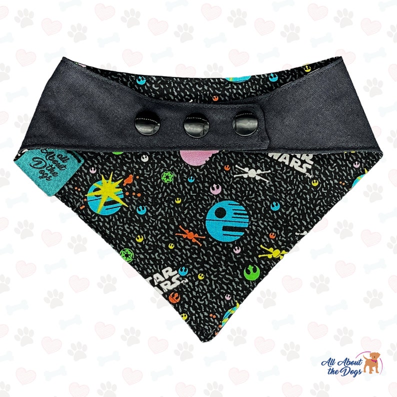 May include: Black and white dog bandana with a Star Wars themed print featuring spaceships, planets, and the words "Star Wars". The bandana has a black snap closure.