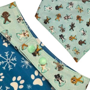 K&ouml;nnte beinhalten: Hunde-Bandana mit blau-wei&szlig;em Schneeflocken- und Pfotenmuster. Der Bandana hat einen hellblauen Besatz mit Hundeengel-Illustrationen und zwei gr&uuml;nen Kn&ouml;pfen zum Verschlie&szlig;en.