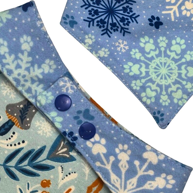 May include: Close-up of two dog bandanas. One is light blue with white snowflakes and paw prints. The other is light blue with a floral and mushroom pattern. Both bandanas have blue snap closures.