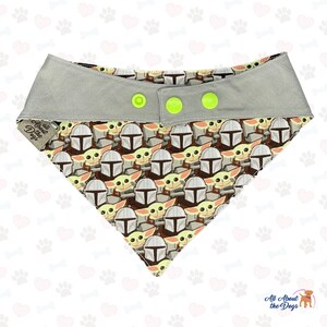 May include: A grey dog bandana with a repeating pattern of a cartoon character wearing a helmet. The bandana has three yellow buttons and the text "All About the Dogs" is printed on the left side.
