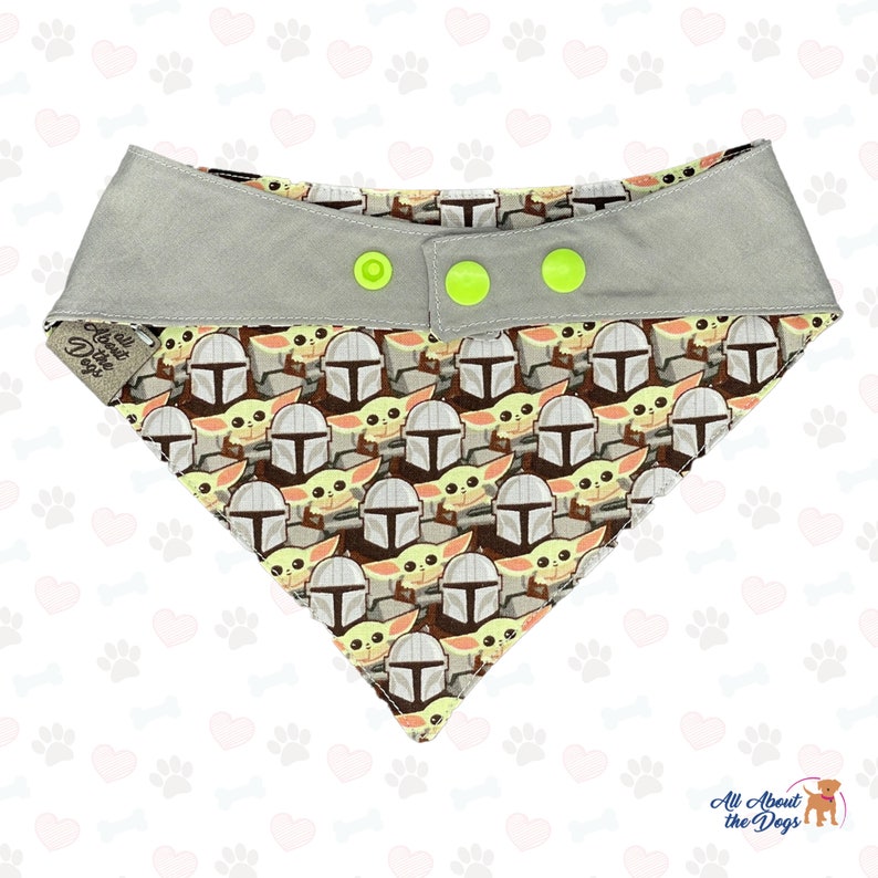 May include: A grey dog bandana with a repeating pattern of a cartoon character wearing a helmet. The bandana has three yellow buttons and the text "All About the Dogs" is printed on the left side.