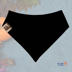 Peut inclure: Bandana noire pour chien, de forme triangulaire. L'accessoire porte une petite &eacute;tiquette avec le texte "All About the Dogs". L'arri&egrave;re-plan comprend une citrouille et des feuilles d'automne.