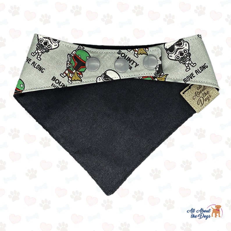 May include: A gray and black dog bandana with a Star Wars theme. The bandana features a repeating pattern of characters from the Star Wars franchise, including Boba Fett, Stormtroopers, and the text "Move Along" and "Bounty Hunter". The bandana has a black fleece lining and snap closures.