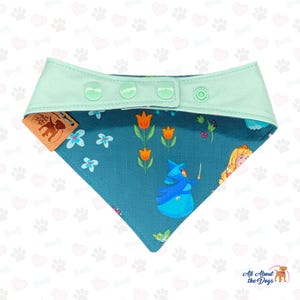 May include: A teal dog bandana with a mint green collar and snap closure. The bandana features a floral and fairy tale design with orange tulips, blue flowers, and a fairy character. A brown tag with a dog silhouette is attached.