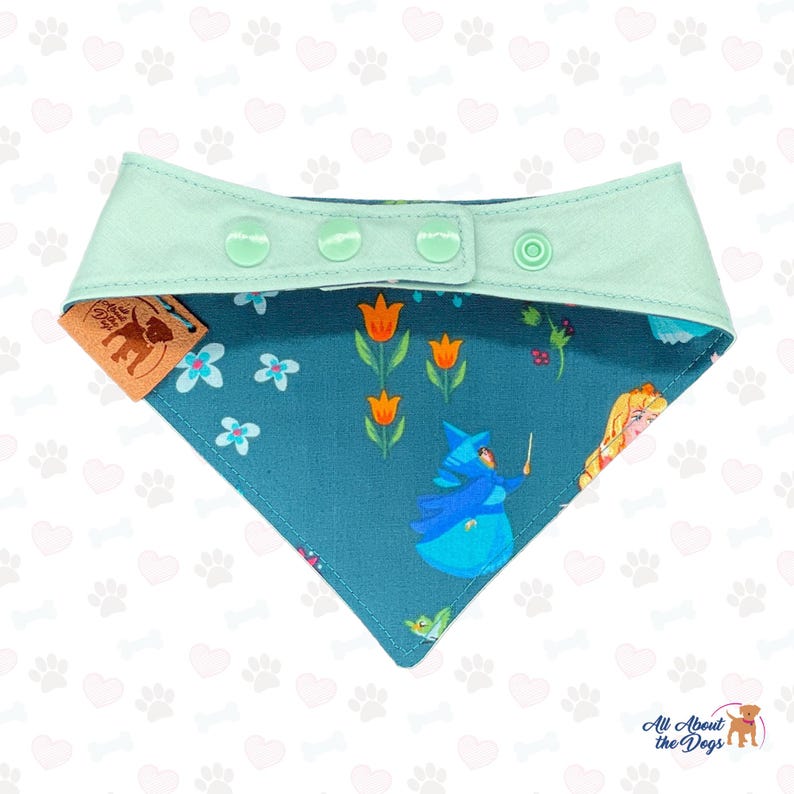 May include: A teal dog bandana with a mint green collar and snap closure. The bandana features a floral and fairy tale design with orange tulips, blue flowers, and a fairy character. A brown tag with a dog silhouette is attached.