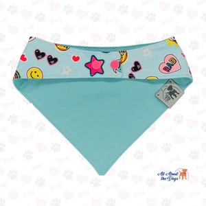 May include: A light blue dog bandana with a patterned top featuring hearts, stars, and smiley faces. The bandana has a triangular shape and a tag with a dog silhouette. The text "RAD" is visible on the top.