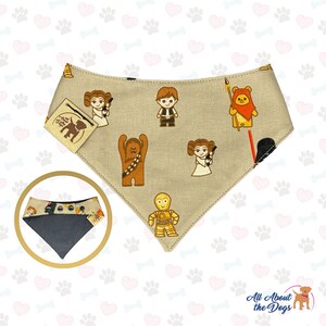 K&ouml;nnte beinhalten: Ein beiger Hundehalstuch mit schwarzem Besatz zeigt ein sich wiederholendes Muster von Star Wars-Charakteren, darunter Prinzessin Leia, Chewbacca, Darth Vader und R2-D2. Das Halstuch hat ein braunes Leder-Etikett mit dem Text "All About the Dogs" und einen Pfotenabdruck.