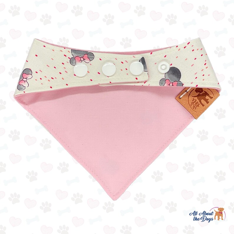 May include: A pink and white dog bandana with a playful design. The top band features a pattern of gray mouse heads with pink bows and red dots. The bandana has three white snap closures and a pink triangle bottom. A brown tag reads "All About the Dogs".