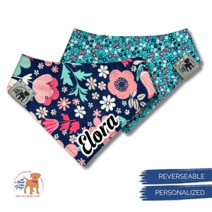 K&ouml;nnte beinhalten: Zwei wendbare Hunde-Bandanas. Eines ist marineblau mit rosa und wei&szlig;en Blumenmustern, das andere ist t&uuml;rkis mit einem kleinen Blumendruck. Der Name "Elora" ist auf das marineblaue Bandana gedruckt. Die Bandanas haben ein Etikett mit einem Hunde-Logo.