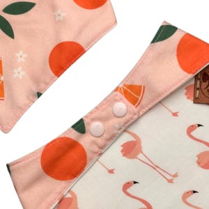 May include: Two baby bibs with a pink and white pattern. One bib features orange fruit and green leaves, while the other has a flamingo design. The bibs have snap closures for easy use.