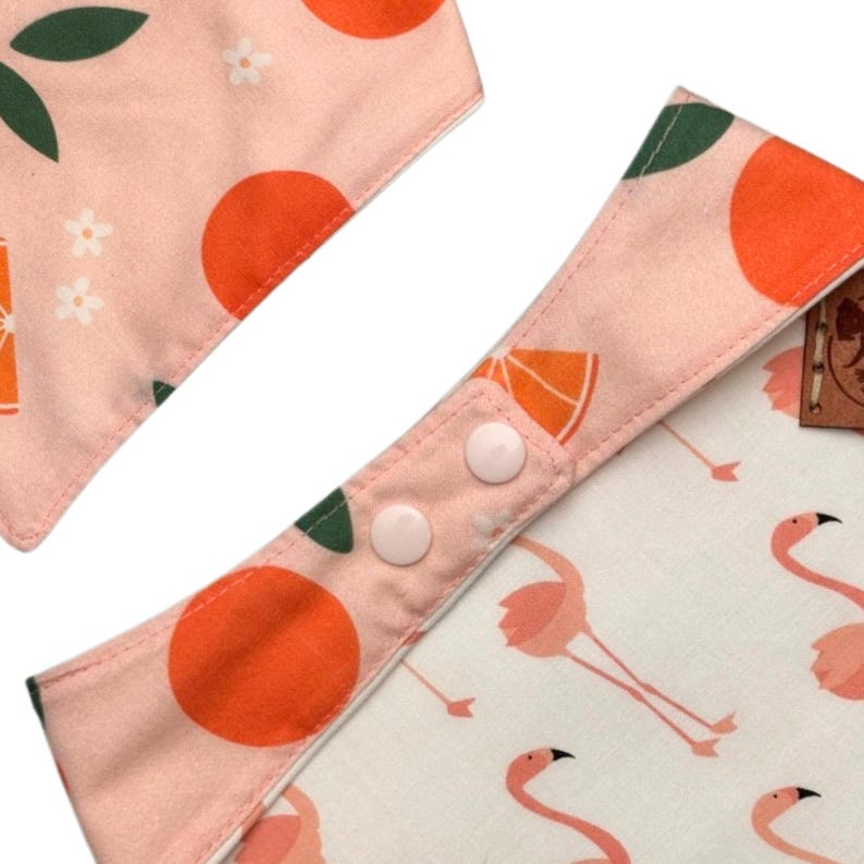 May include: Two baby bibs with a pink and white pattern. One bib features orange fruit and green leaves, while the other has a flamingo design. The bibs have snap closures for easy use.