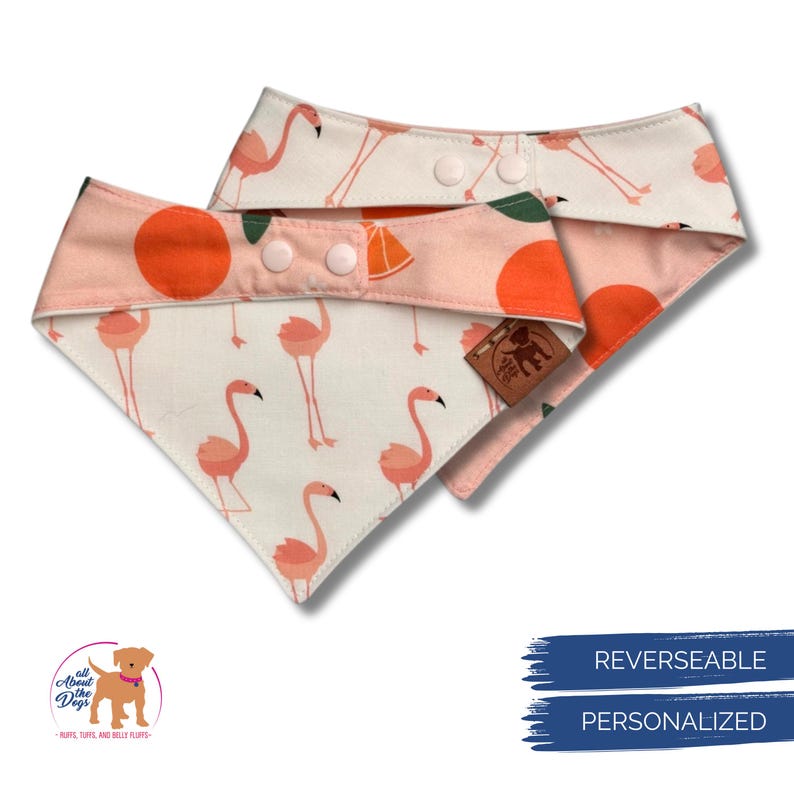May include: Two reversible dog bandanas. One features a flamingo pattern on a white background, the other has an orange slice pattern. The bandanas are secured with pink snaps. The logo says "All About the Dogs".